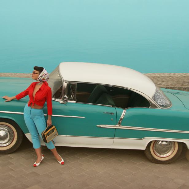 Eine Pin-up-Frau posiert mit einem Radio neben einem türkisfarbenen Oldtimer.