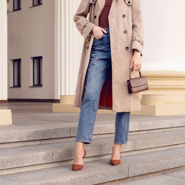 Auf dem Bild ist eine Frau zu sehen, die modisch gekleidet ist mit Jean, Trenchcoat und Tasche in der Hand.