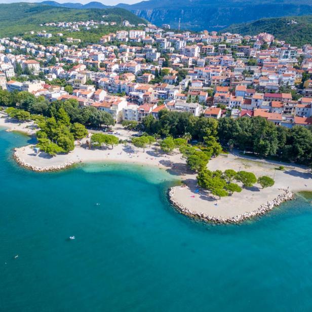 Luftaufnahme der Kvarner Bucht in Kroatien mit türkisblauem Meer, feinem Strand, grünen Pinienbäumen und malerischer Küstenstadt mit roten Ziegeldächern im Hintergrund.