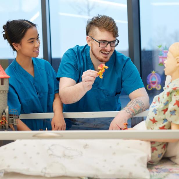 Zwei Pfleger:innen bespaßen eine Puppe in einem Krankenbett. 