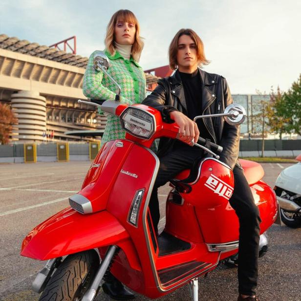 Ein Mann und eine Frau posieren auf einem roten Lambretta-Roller vor einem Stadion.