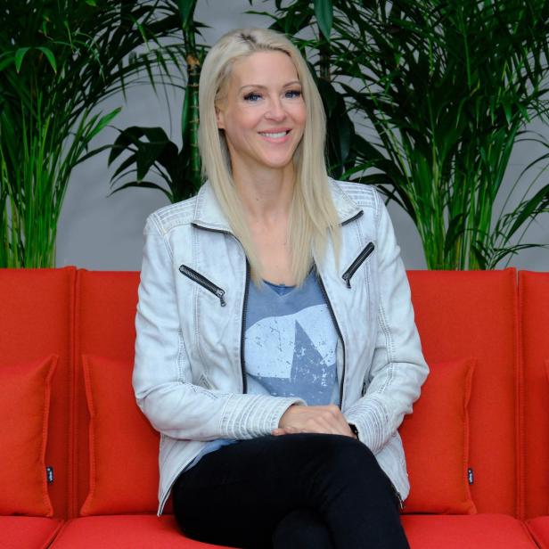 Simone Lugner sitzt auf einem roten Sofa vor grünen Pflanzen.