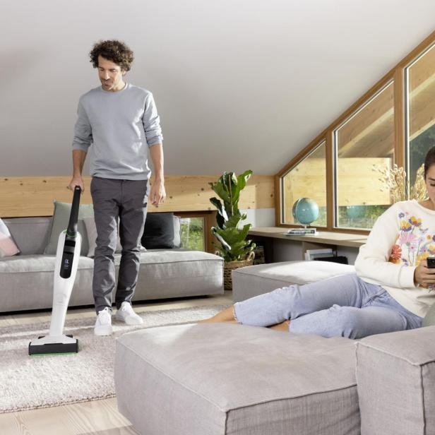 Eine Frau sitzt auf der Couch im Wohnzimmer und tippt auf ihrem Handy, während der Mann den Teppich staubsaugt.