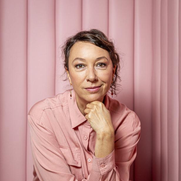 Ursula Strauss sitzt vor einem rosa Hintergrund.