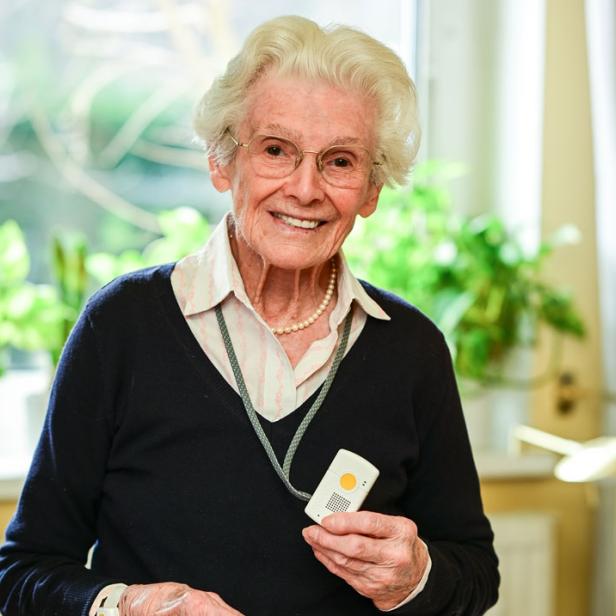 Eine ältere Dame mit einem mobilen Notrufgerät lächelt in die Kamera.