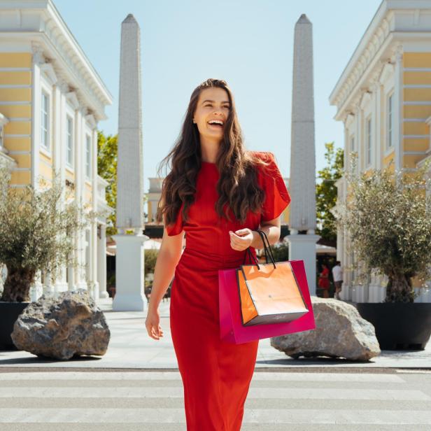 Eine Frau in einem roten Kleid hält mehrere Einkaufssackerl in der Hand und lächelt. Im Hintergrund ist ein Shoppingcenter zu sehen.