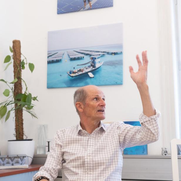Wolfang Putschek von Swimsol sitzt vor einer Wand und bewegt eine Hand in der Luft, während er etwas erklärt. Hinter ihm ist ein Bild von den Swimsol-Anlagen am Meer