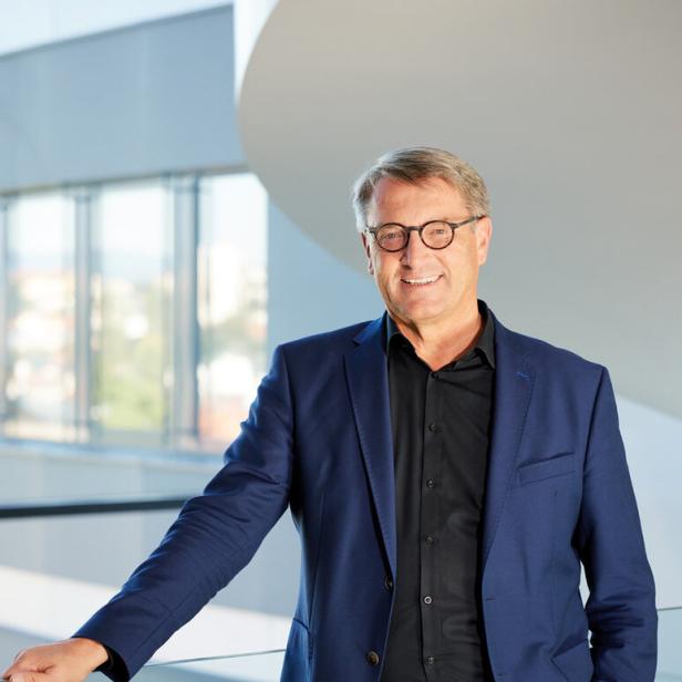 Ein lächelnder Mann mit Brille im blauen Sakko steht an einem Geländer in einem hellen Innenraum.