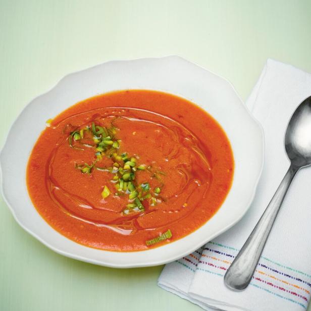 Ein Teller Gazpacho, garniert mit gehackten Kräutern und einem Schuss Öl, steht neben einem Löffel.