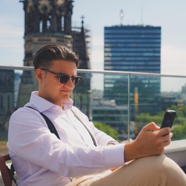 Junger Mann mit Smartphone sitzt auf einer Dachterrasse