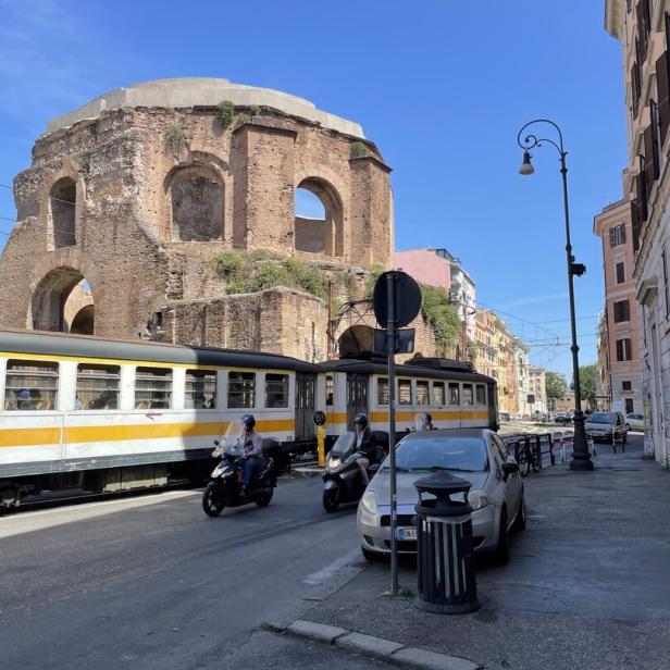 Eine Straße in Rom mit einer alten Ruine, einer Straßenbahn und Rollern.