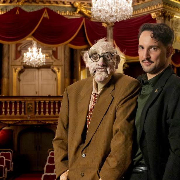 Zwei Männer stehen vor einem großen Theatersaal und blicken in die Kamera.
