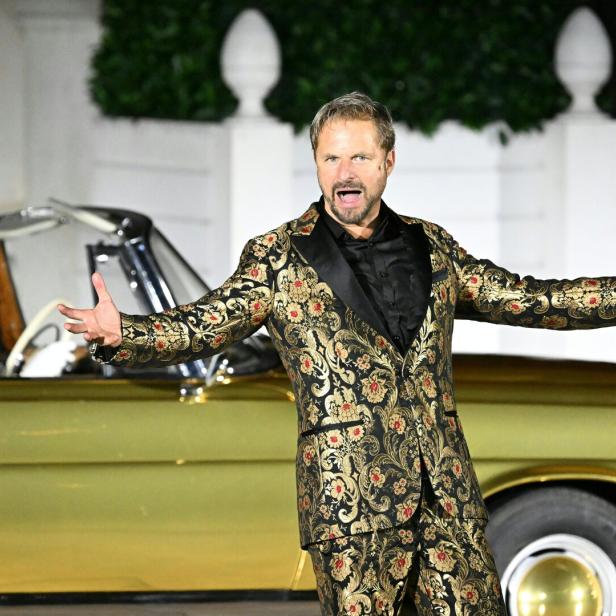 Ein Mann in einem auffälligen, gold-schwarz gemusterten Anzug gestikuliert vor einem goldenen Oldtimer mit Chauffeur.