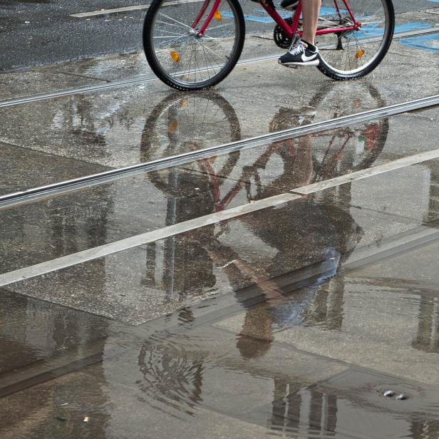 Ein Radfahrer fährt bei Regen über eine von Pfützen bedeckte Straße, in denen sich die Umgebung spiegelt.