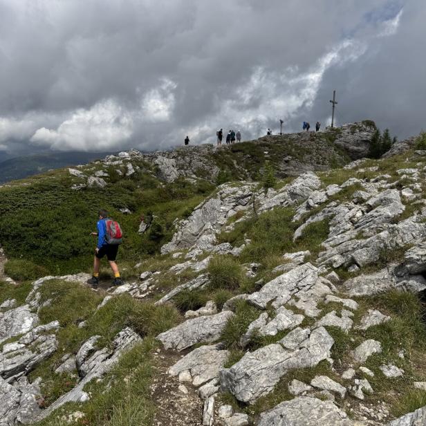 Ein Wanderer steigt einen felsigen Pfad auf einen Berggipfel hinauf, wo sich weitere Personen aufhalten.