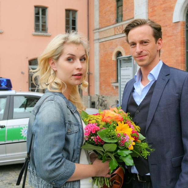 Eine Frau mit Blumenstrauß und ein Mann stehen vor einem Polizeiauto und einem Gebäude mit Backsteinfassade.