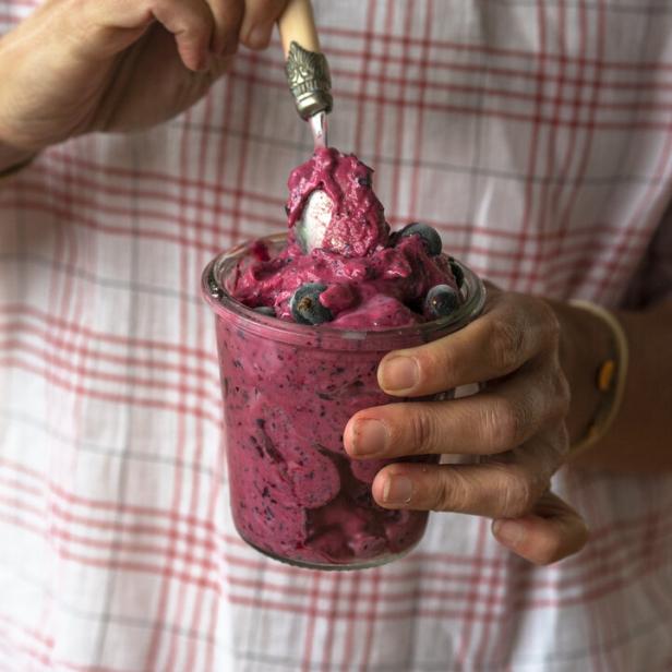 Eine Person hält ein Glas mit rosa Eis und Blaubeeren in der Hand.