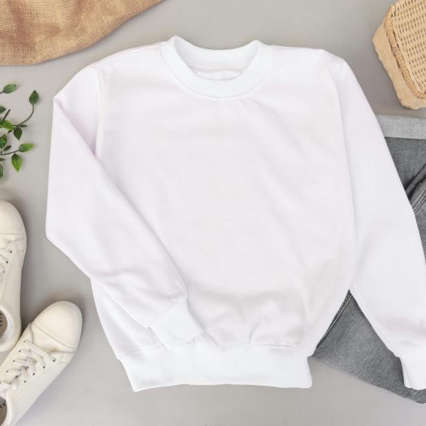 Ein weißer Pullover liegt schon aufgelegt auf dem Boden, daneben eine graue Jeans, weiße Sneaker und eine Korbtasche.