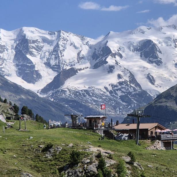 Blick auf ein Bergrestaurant mit Spielplatz vor einer schneebedeckten Bergkette in den Alpen.