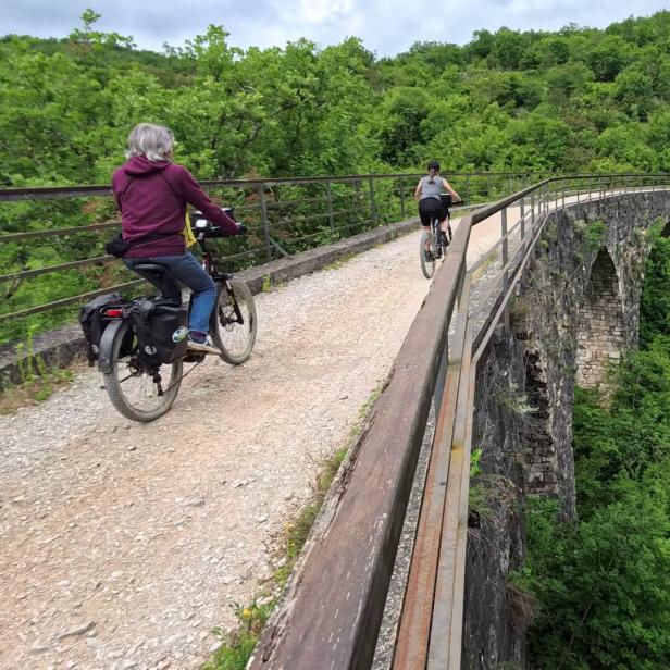 Zwei Personen fahren mit Fahrrädern über eine alte Steinbrücke in einer grünen Landschaft.