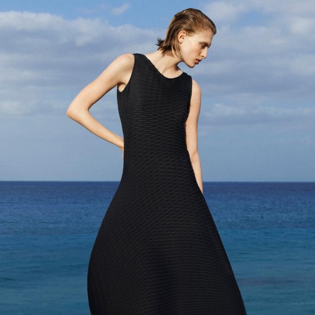 Eine Frau in einem schwarzen Kleid und Sandalen steht vor dem Meer.