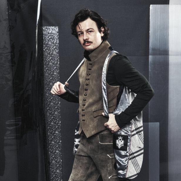 Ein Mann mit Schnurrbart trägt eine Lederhose, eine Weste und Stiefel vor einem grauen Hintergrund.