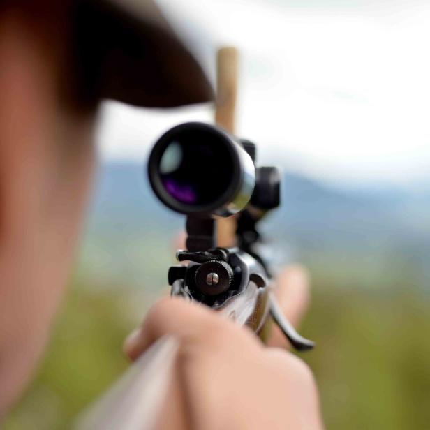 Ein Jäger zielt mit einem Gewehr samt Zielfernrohr auf eine unscharfe Landschaft.