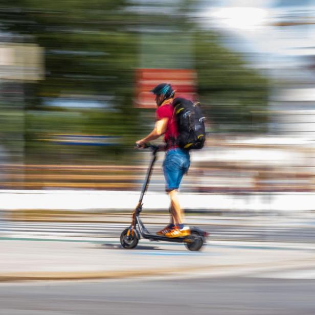 Ein Mann mit Rucksack fährt mit einem Elektroroller über einen Zebrastreifen.