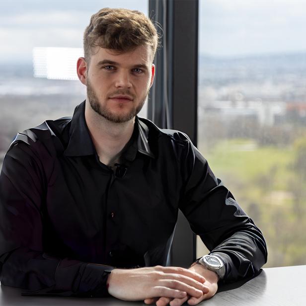 Ein Mann in einem schwarzen Hemd, der an einem Konferenztisch vor einem Fenster sitzt. 
