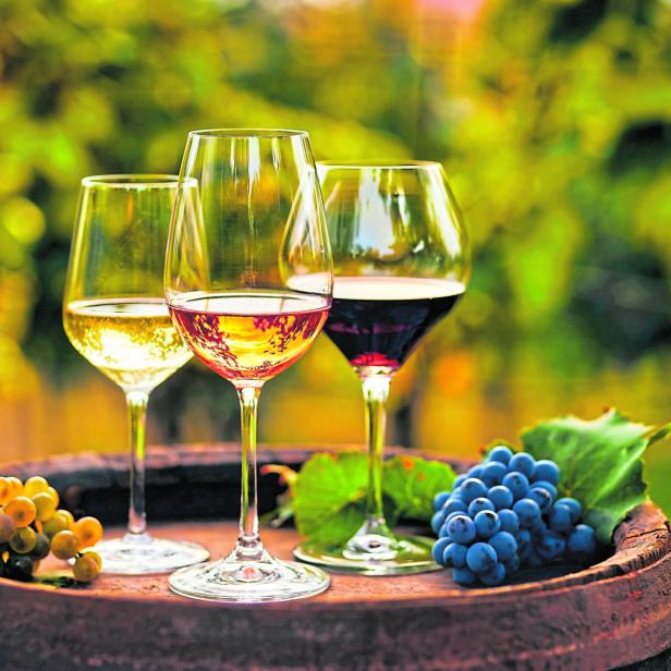 Drei Gläser Weiß-, Rosé- und Rotwein stehen auf einem alten Holzfass mit Weintrauben.
