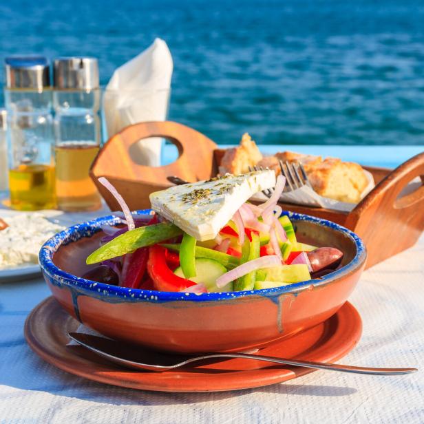 Ein griechischer Salat und Tsatsiki stehen auf einem Tisch einer Taverne in Griechenland mit Blick auf das Meer.
