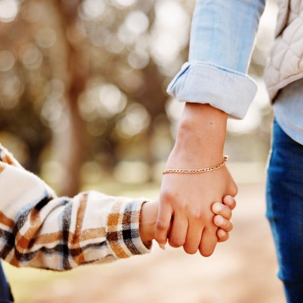Eine Mutter und ihr Kind halten Händchen und gehen mit einem Schulrucksack durch den Park.