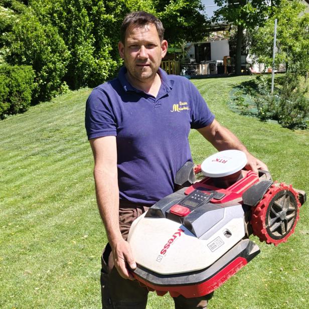 Ein Mann steht mit einem Roboter-Rasenmäher in der Hand in einem Garten.