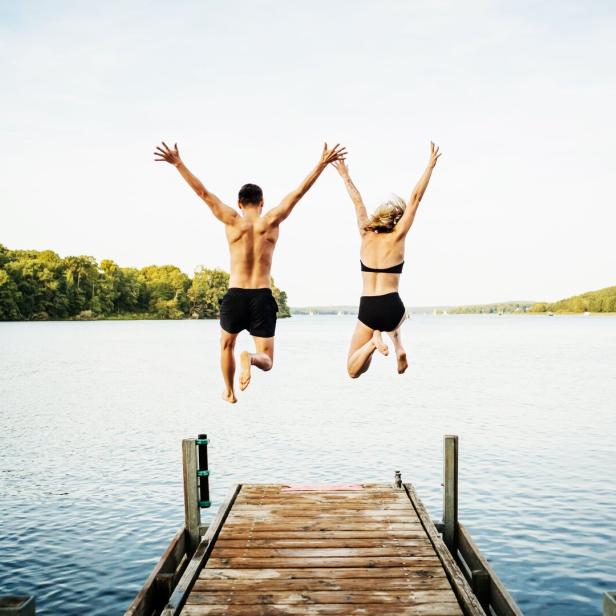 Two Friends Jumping Off Jetty At Lake Together