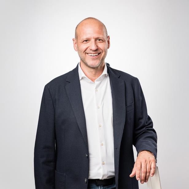 Ein Mann steht mit Sakko und weißem Hemd vor einem grauen Hintergrund und schaut in die Kamera.