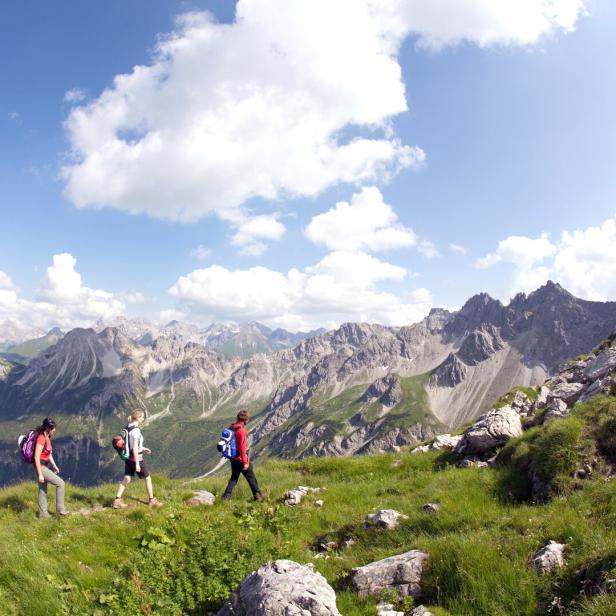 Drei Wanderer gehen auf einem grasbewachsenen Bergrücken mit Blick auf eine Bergkette.