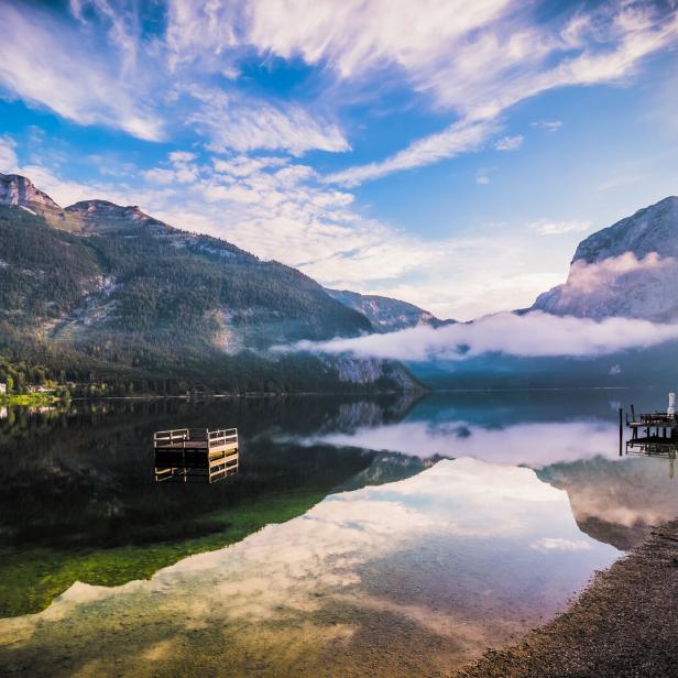 Breathtaking morning view of a woman standing on the shore of Altausseer See (Lake Aussee), Styria, Austria, drinking coffee and looking at the lake