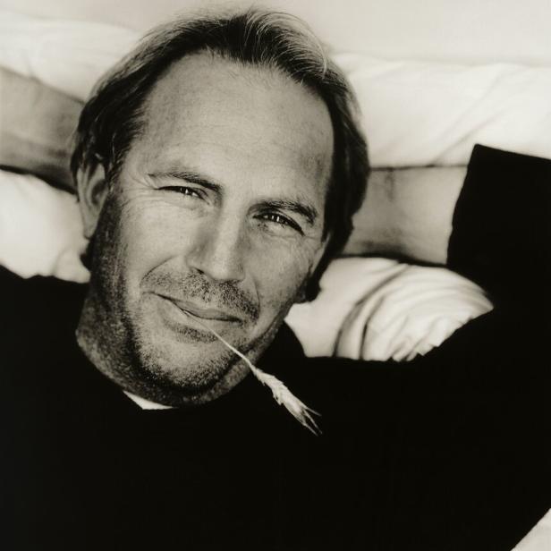 Ein Schwarzweißporträt von Kevin Costner mit einem Grashalm im Mund.