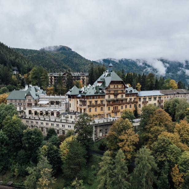 Das große Südbahnhotel mit seinen verschiedenen Trakten und großem Balkon. Es thront auf einem bewaldeten Hang. Im Hintergrund erheben sich Berge, Nebel lichtet sich.