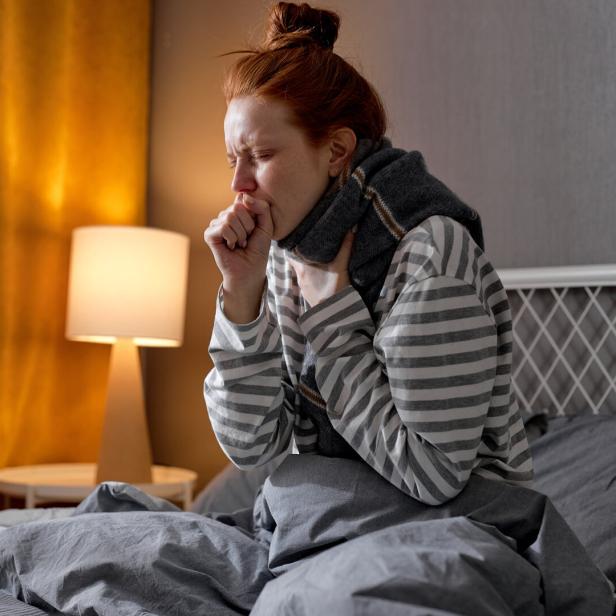 Frau ist krank und sitzt hustend im Bett.