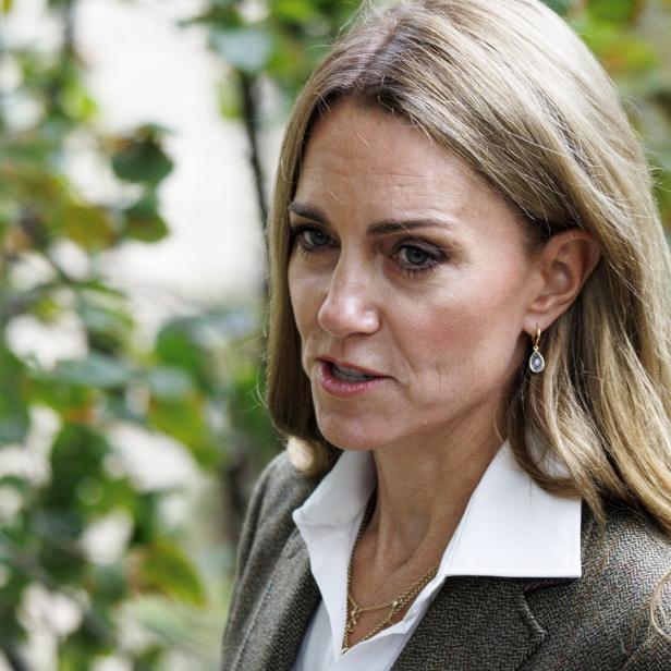Prinzessin Kate steht draußen vor grünem Laub. Sie trägt ein Sakko mit weißem Hemd und Ohrringe.
