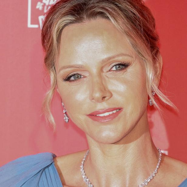 Fürstin Charlène von Monaco trägt ein blaues Kleid und eine funkelnde Halskette vor einem roten Hintergrund.