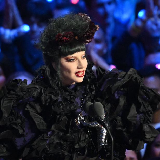 Eine Sängerin in einem extravaganten schwarzen Kleid steht auf einer Bühne vor Publikum.