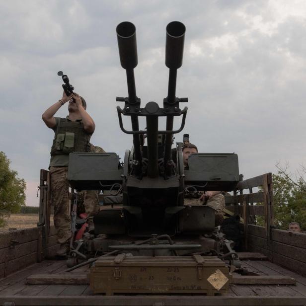 Zwei Soldaten stehen auf einem Militärfahrzeug mit einer großen Flugabwehrkanone, einer blickt durch ein Fernglas in den Himmel.