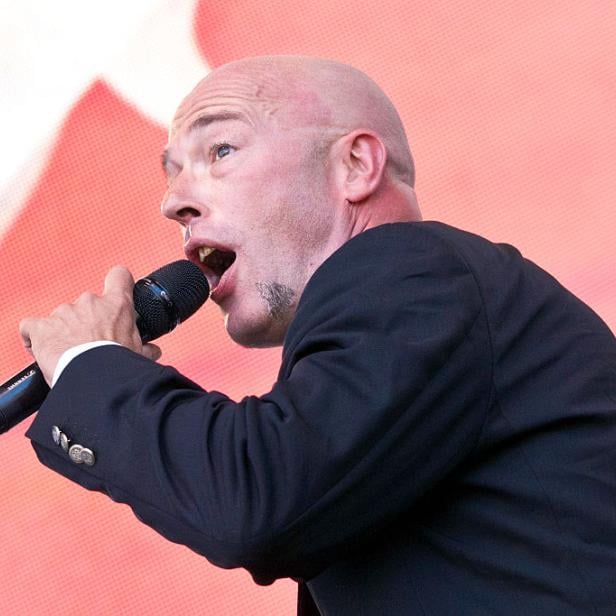 Der Graf singt auf der Bühne in ein Mikrofon, das er in der Hand Hält. Er steht vor einem rotem Hintergrund.