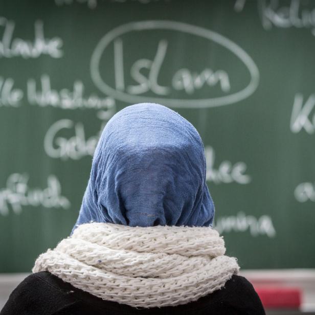 Eine Person mit blauem Kopftuch und weißem Schal steht vor einer Tafel mit islambezogenen Begriffen.