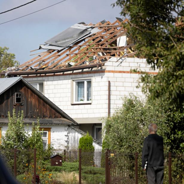 Ein Haus mit stark beschädigtem Dach, bei dem große Teile der Dacheindeckung fehlen, steht inmitten von Bäumen und Sträuchern.