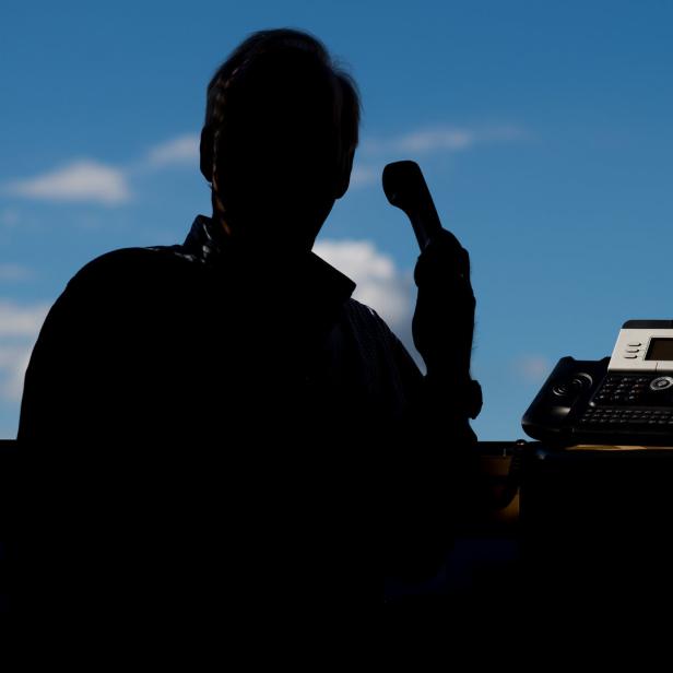 Eine Silhouette einer Person telefoniert mit einem Tischtelefon vor einem Fenster mit blauem Himmel.