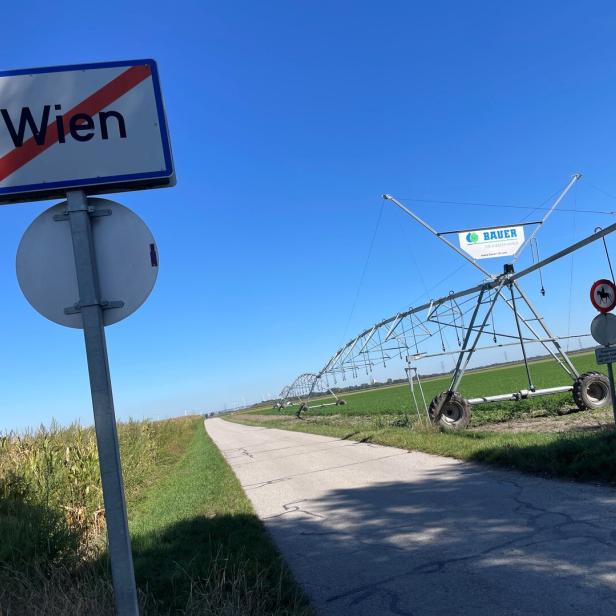 Ein Ortsausgangsschild mit „Wien“ steht an einer Landstraße neben Feldern und einer großen landwirtschaftlichen Bewässerungsanlage.
