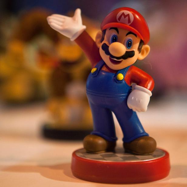 Eine Mario-Figur steht auf einem Schachbrettmuster-Sockel und winkt.